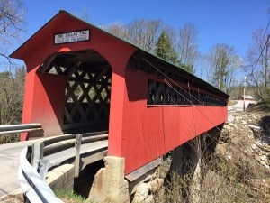 Red painted covered bridge with one lane road surrounded by trees and bright blue skies.