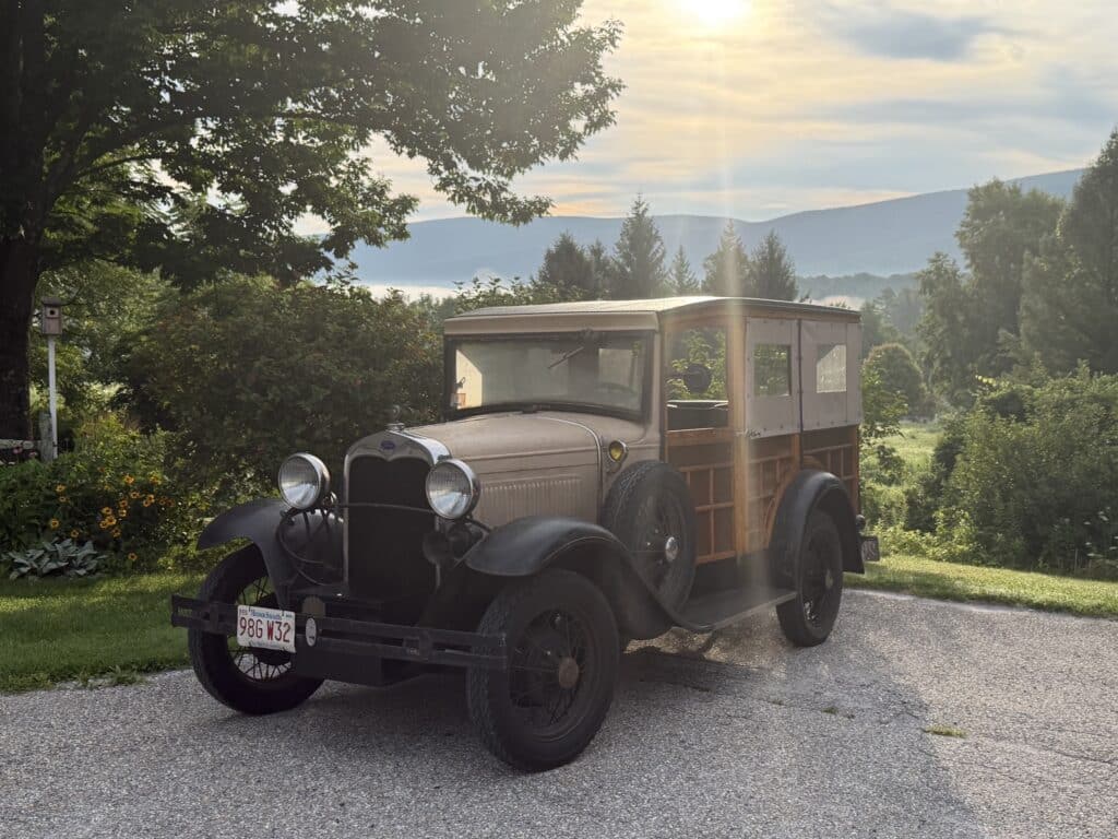 Vintage wood-sided pickup truck parked on a gravel driveway, garden in bloom with hills beyond.