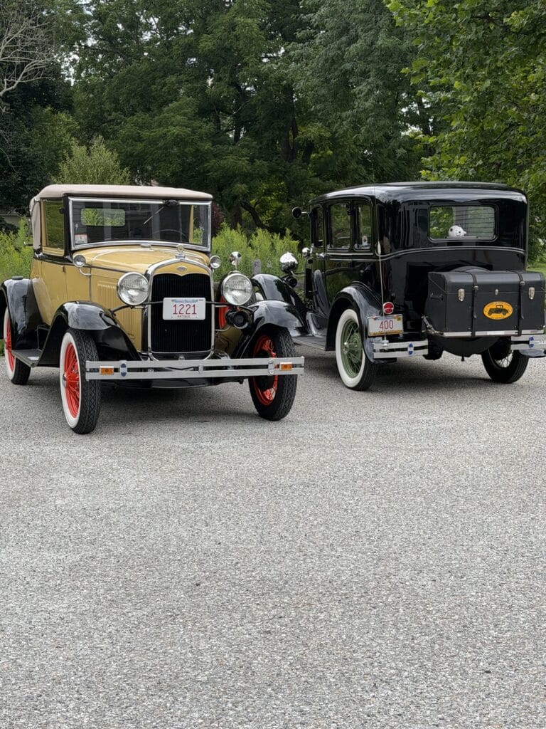 Two classic vintage cars parked on a paved road, beige/yellow on the left with red wheels and a glossy black car on the right, greenery behind them.