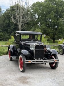 Vintage black car with red wheels parked on a driveway, man visible in the driver's seat, greenery in the background.