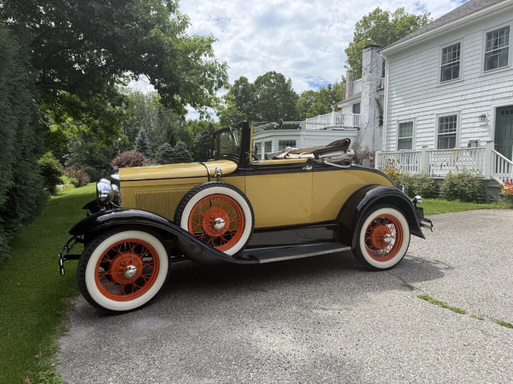 Vintage yellow convertible with orange-spoked wheels parked on a driveway beside a white house and green trees.