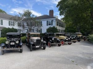 Row of vintage antique cars parked in front of a white house with green shutters and trees nearby, sunny day