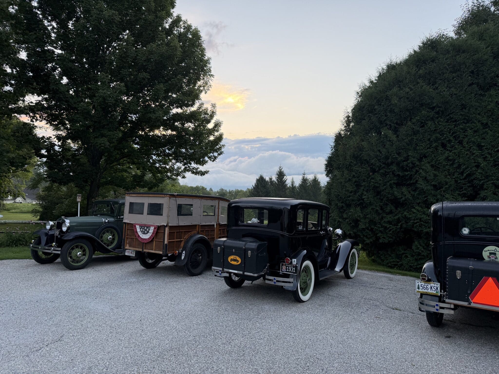 Row of vintage cars and a wooden wagon parked in a gravel lot, with green trees and a sunset sky in the background.