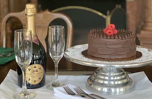 A white place setting with champaigne flutes, and champaigne bottle, next to a cake service and chocolate iced cake with raspberries on top.