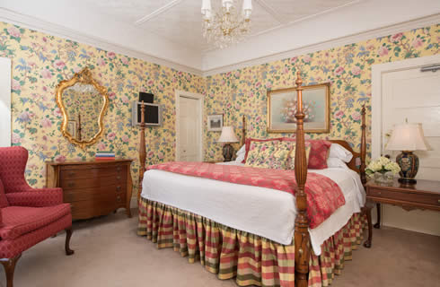 Bedroom with yellow chintz wallpaper, a four-post bed, antique wooden furniture and a red wing back chair.