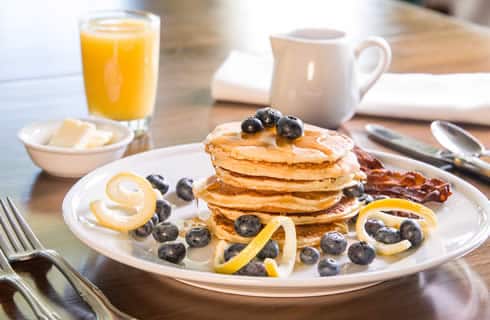 A stack of fluffy pancakes garnished with blueberries accompanies bacon on a white plate next to syrup and juice.