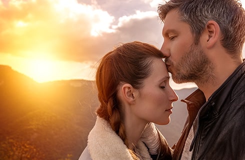 Sun setting behind a mountain between clouds in the background as a man kisses a woman on the forehead.