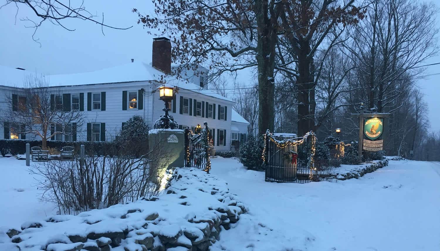 Large white house with black roof and shutters in wintertime with snow and bare trees. 