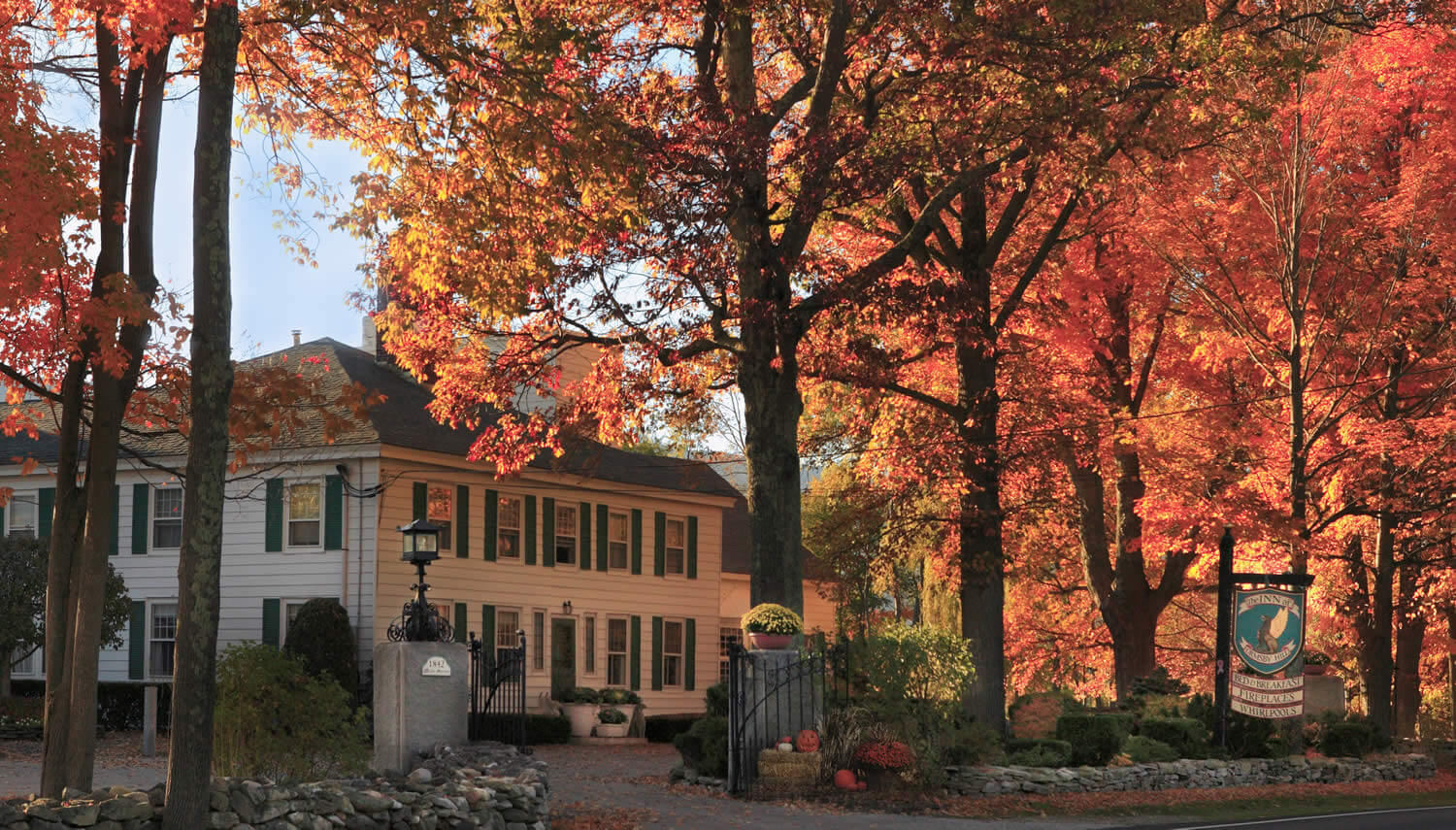 Large white house with black roof and shutters set amid glowing fall foliage. 