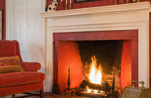 Plaster fireplace with a white mantle next to a red velvet chair in a room with red walls and white trim.