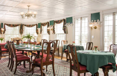 Bright dining rooom with large windows and various tables covered in green cloths set with napkins and silver.