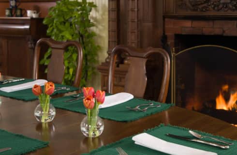 A wooden table set with green placemats and white napkins with silver and tulips in vases next to a fireplace.