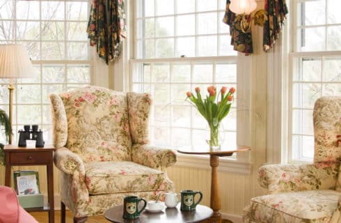 Charming sitting area with two cream and rose wingback chairs, a table with two mugs and large bright windows.