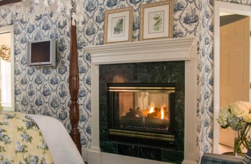 Fireplace with tiled face and white wood mantel in a bedroom with blue patterned wallpaper.