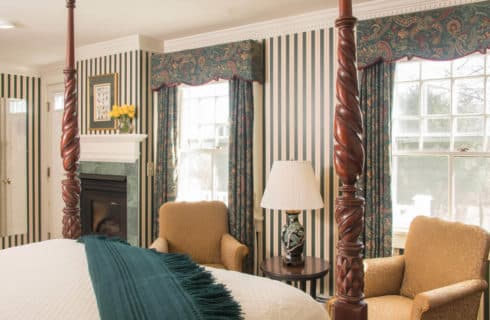 Charming room with a four-post bed, two chairs, green striped wallpaper, a fireplace faced in green marble and two large windows.