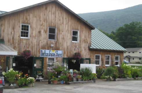 Wooden barn building surrounded with plants and flowers with a sign that says Dutton Farm Stand.