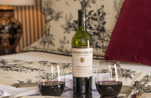 A wine bottle and two stemless glasses on a tray lying on a bed.