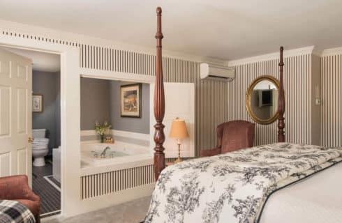 A large four post bed faces a bathroom with a soaking tub and grey tiled floor.