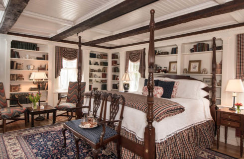 Large timber-ceiling bedroom with an ornate four-post bed, built in library shelbves and a seating area with a table.
