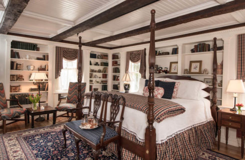 Large timber-ceiling bedroom with an ornate four-post bed, built in library shelbves and a seating area with a table.