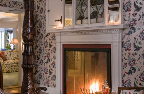 Glass enclosed fireplace in a bedroom with chintz wallpaper and an ornate four-post bed.