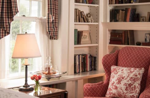 Corner of a bright bedroom with built in library shelves, a red wingback chair and a large window next to the bed.