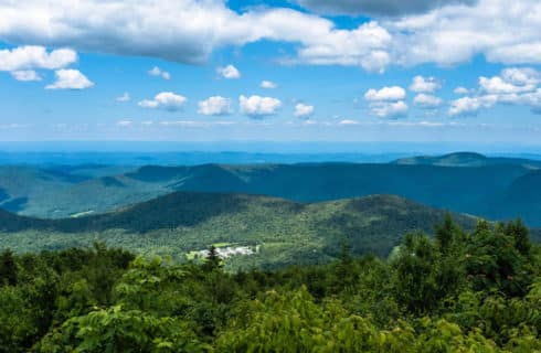 Expansive wide view of tree-covered rolling hills and blue sky with a building right in the middle of it.