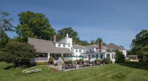 Outdoor back view of the white shingled inn with covered porch, wide center stairs with railing, large stone patio with 3 large outdoor tables with 4 ratan arm chairs each and three kelly green umbrellas with 3 sets of natural wood adironack chair and table vignettes.