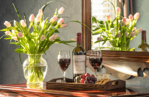 A tray with wine and grapes next to a vase of pink tulips sits on a wooden table below a mirror.