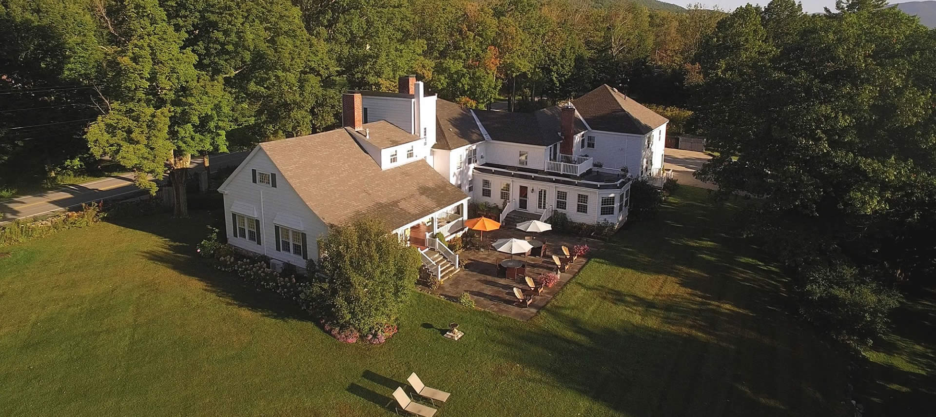 Large white house with a patio and expansive lawn set amongst mature green trees.