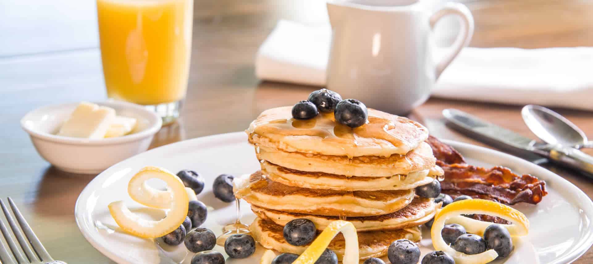 A stack of fluffy pancakes garnished with blueberries accompanies bacon on a white plate next to syrup and juice.