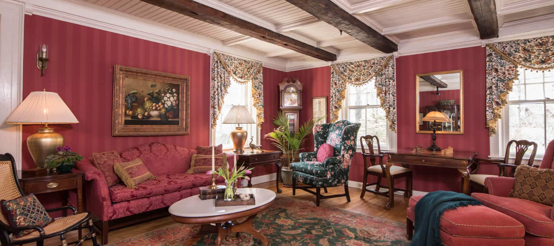 Attractive sitting room papered in red with a sofa, comfortable chairs and a small table desk.