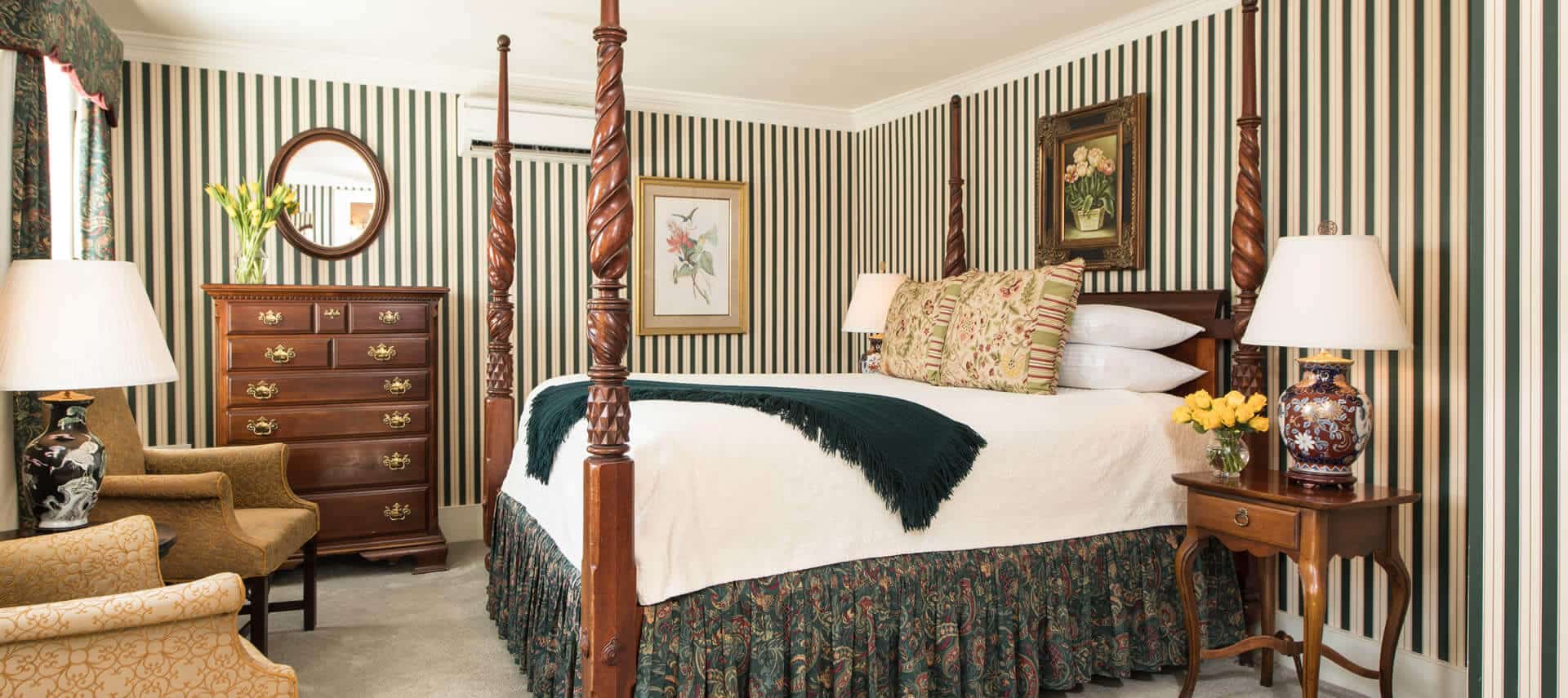 Charming room with a four-post bed, two chairs, green striped wallpaper, and elegant antique wooden furniture.