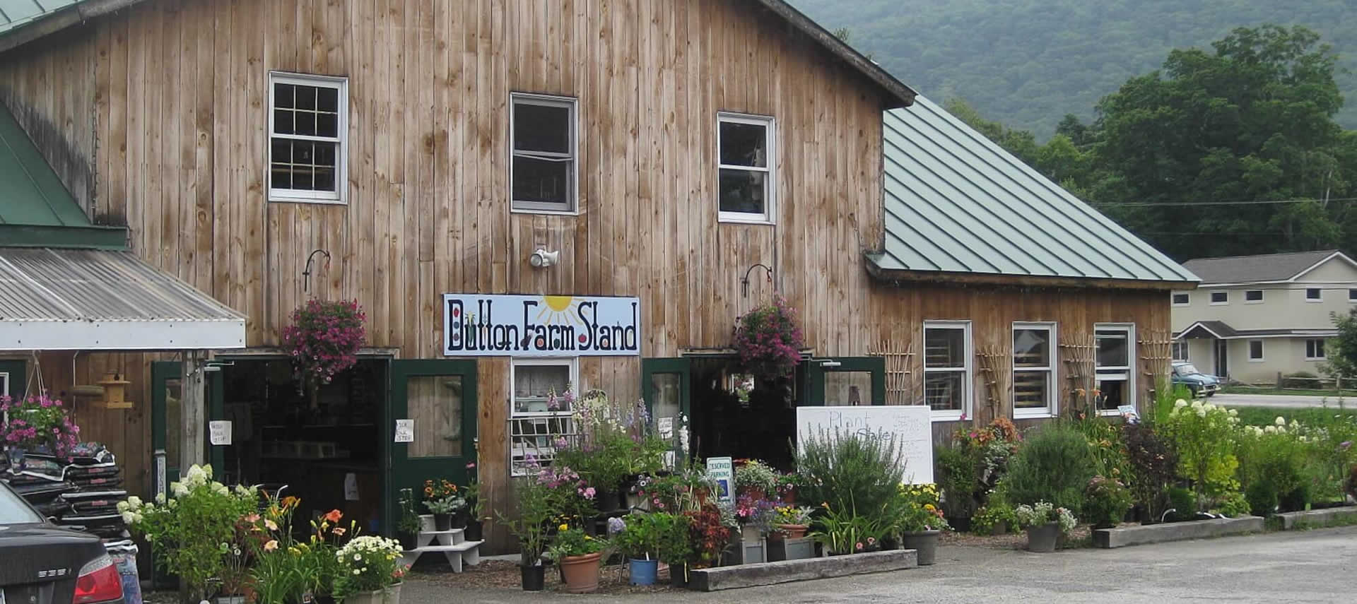 Wooden barn building surrounded with plants and flowers with a sign that says Dutton Farm Stand.