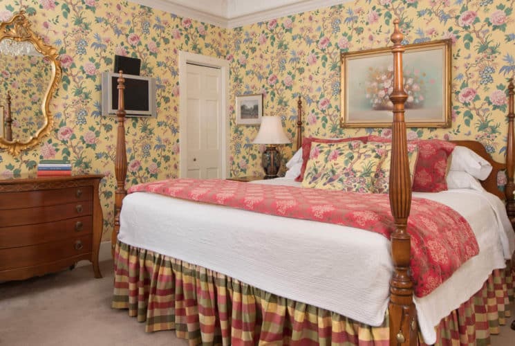 Bedroom with yellow chintz wallpaper, a four-post bed, antique wooden furniture and a red wing back chair.