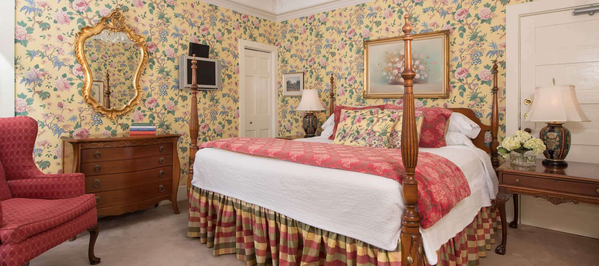 Bedroom with yellow chintz wallpaper, a four-post bed, antique wooden furniture and a red wing back chair.