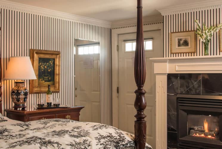 Bedroom with striped wallpaper, a fireplace and a four-post bed with a black and white patterned cover.
