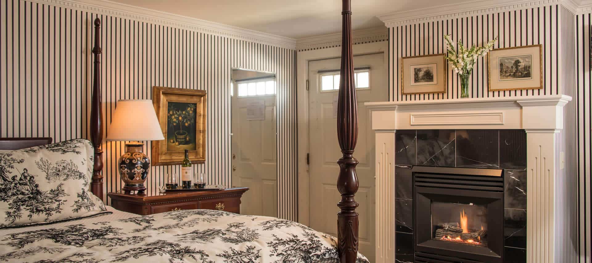 Bedroom with striped wallpaper, a fireplace and a four-post bed with a black and white patterned cover.