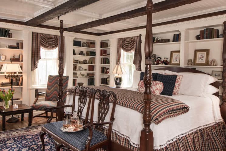 Ornate four poster bed made up in white next to a window and built-in bookcases with a red wingback chair.