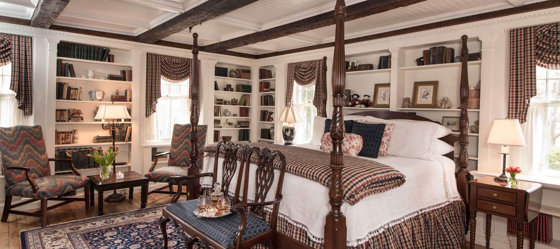 Ornate four poster bed made up in white next to a window and built-in bookcases with a red wingback chair.