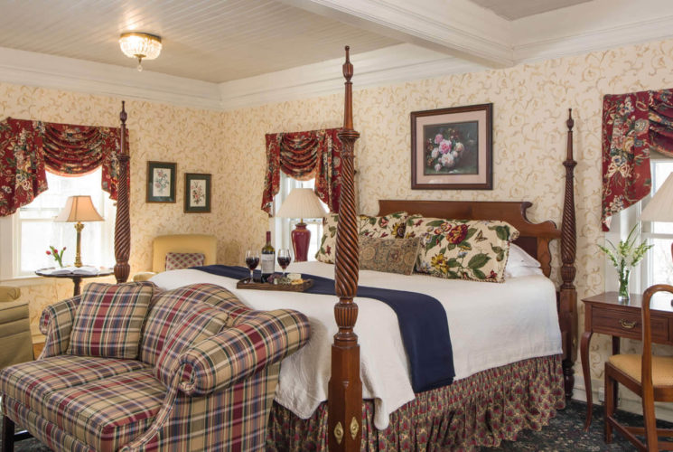 Large bright bedroom with a tall four-post bed, a plaid sofa, a beige armchair and three large windows.