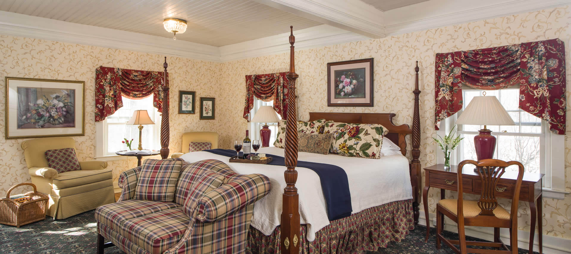 Large bright bedroom with a tall four-post bed, a plaid sofa, a beige armchair and three large windows.