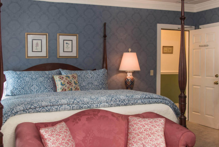 A room with blue wallpaper, a four-post bed with blue coverings, a rose damask sofa and a small wooden desk.