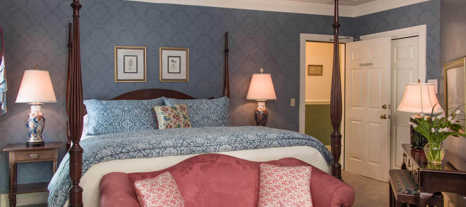 A room with blue wallpaper, a four-post bed with blue coverings, a rose damask sofa and a small wooden desk.