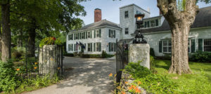 Colonial white house with dark green shutters, large circular driveway with stone gates with gas lamps and wrought iron gates open.