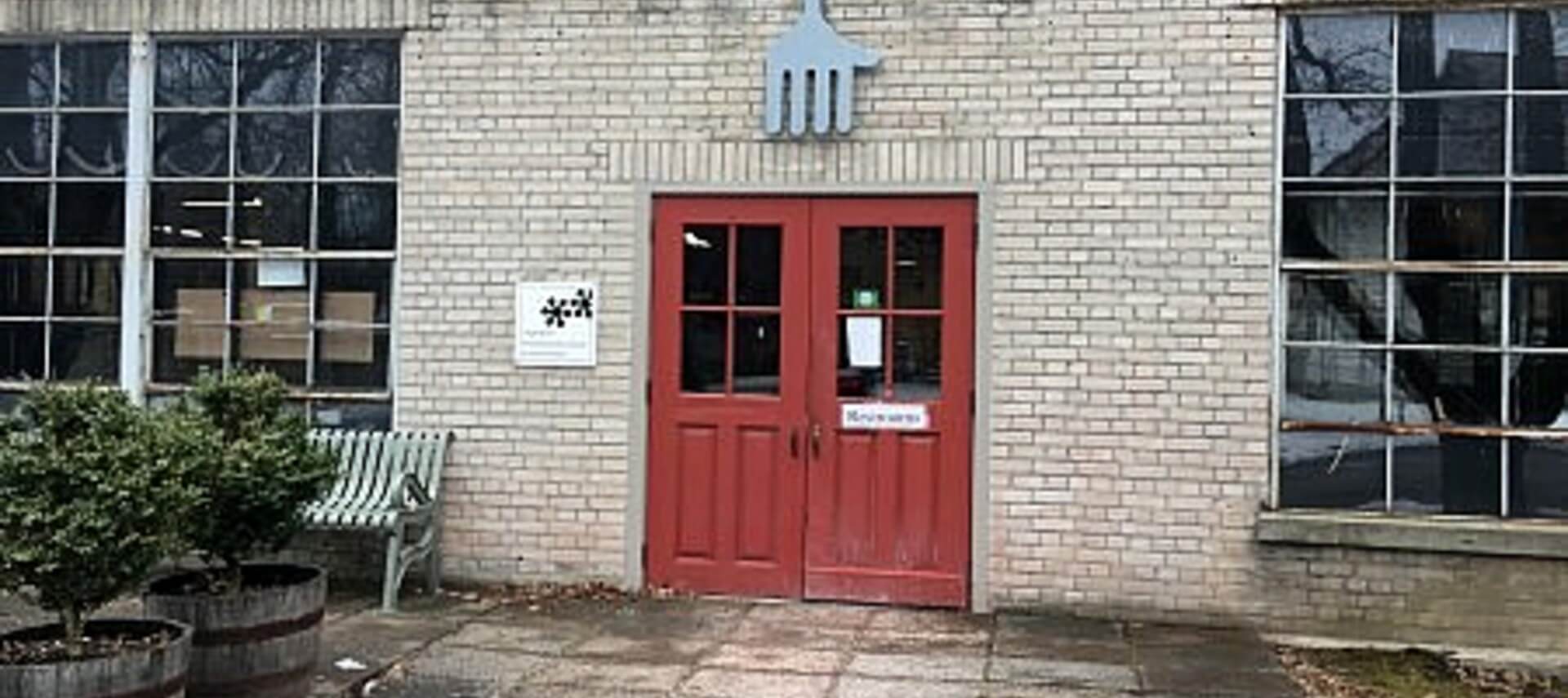 Front facade of cream colored brick building with double red doors in center with windows, flanked by larger single paned windows on each side with metal fork sige above, concrete patio in front and green shrubs in wine barrels.