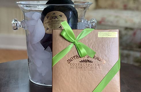 A clear ice bucket chilling a bottle with a brown square package with green ribbon wrapping it diagonally on a black table surface with love seat in background.