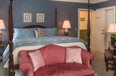 Bedroom with blue wallpaper, a four-post bed and vintage rose colored setee in front.